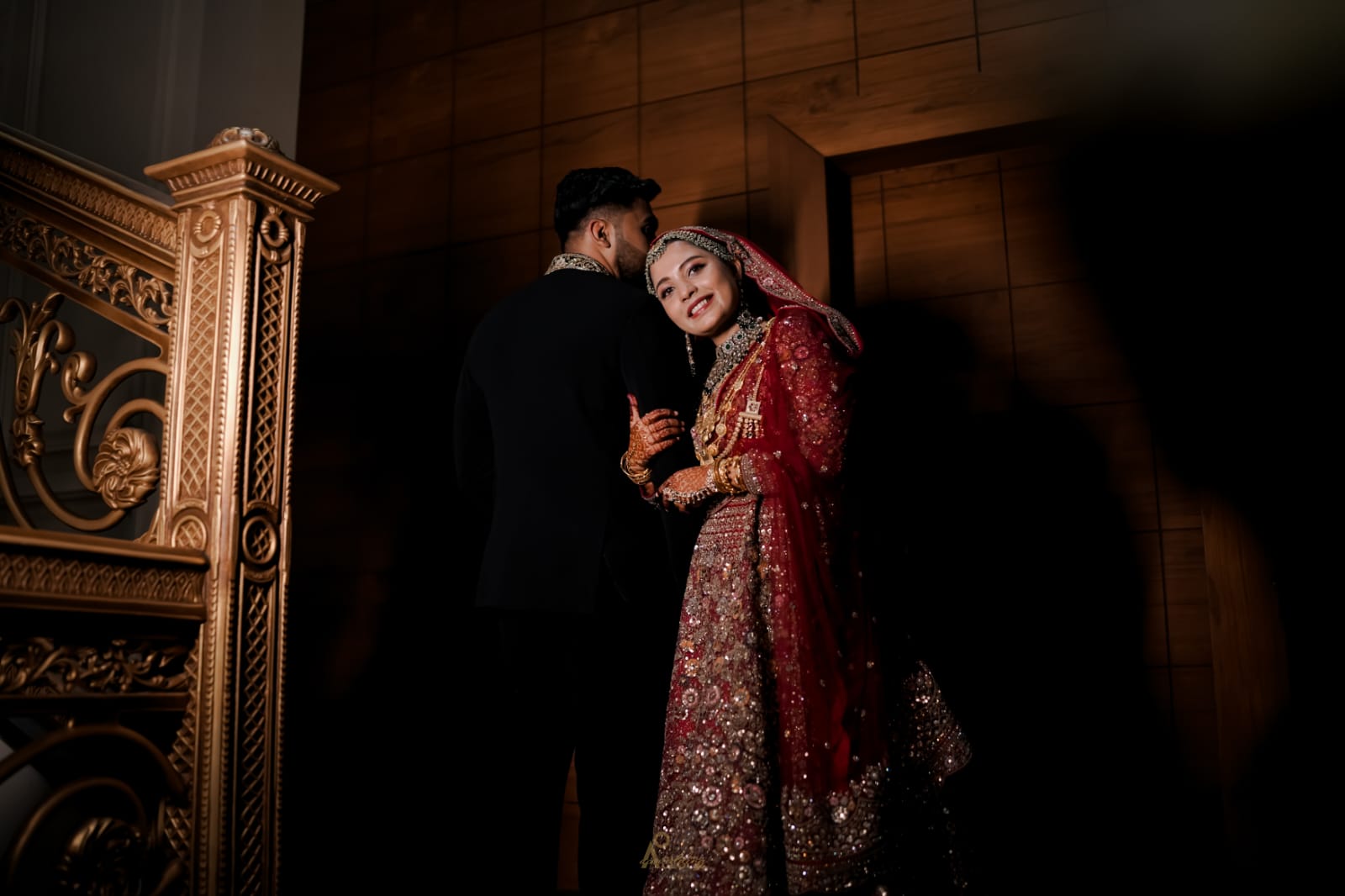 A bride and groom in traditional attire stand closely together in an ornate indoor setting with dramatic golden lighting and an intricately carved pillar in the background.