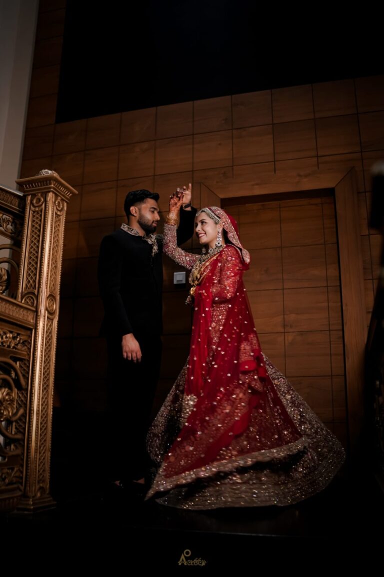 A bride in a traditional red and gold lehenga twirls while holding hands with a groom dressed in black during an indoor wedding ceremony, with ornate golden décor and wooden panel walls in the background