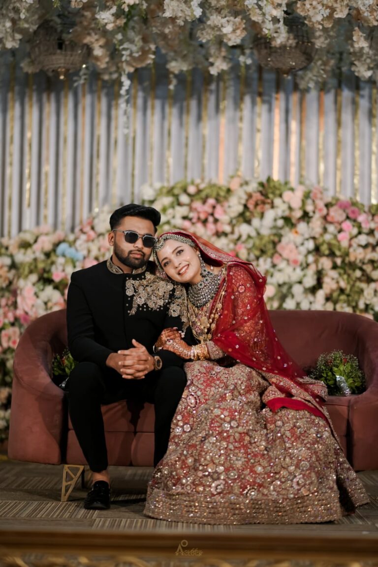 A wedding couple sitting on a sofa, with the bride dressed in traditional attire and the groom in a suit, surrounded by floral decorations