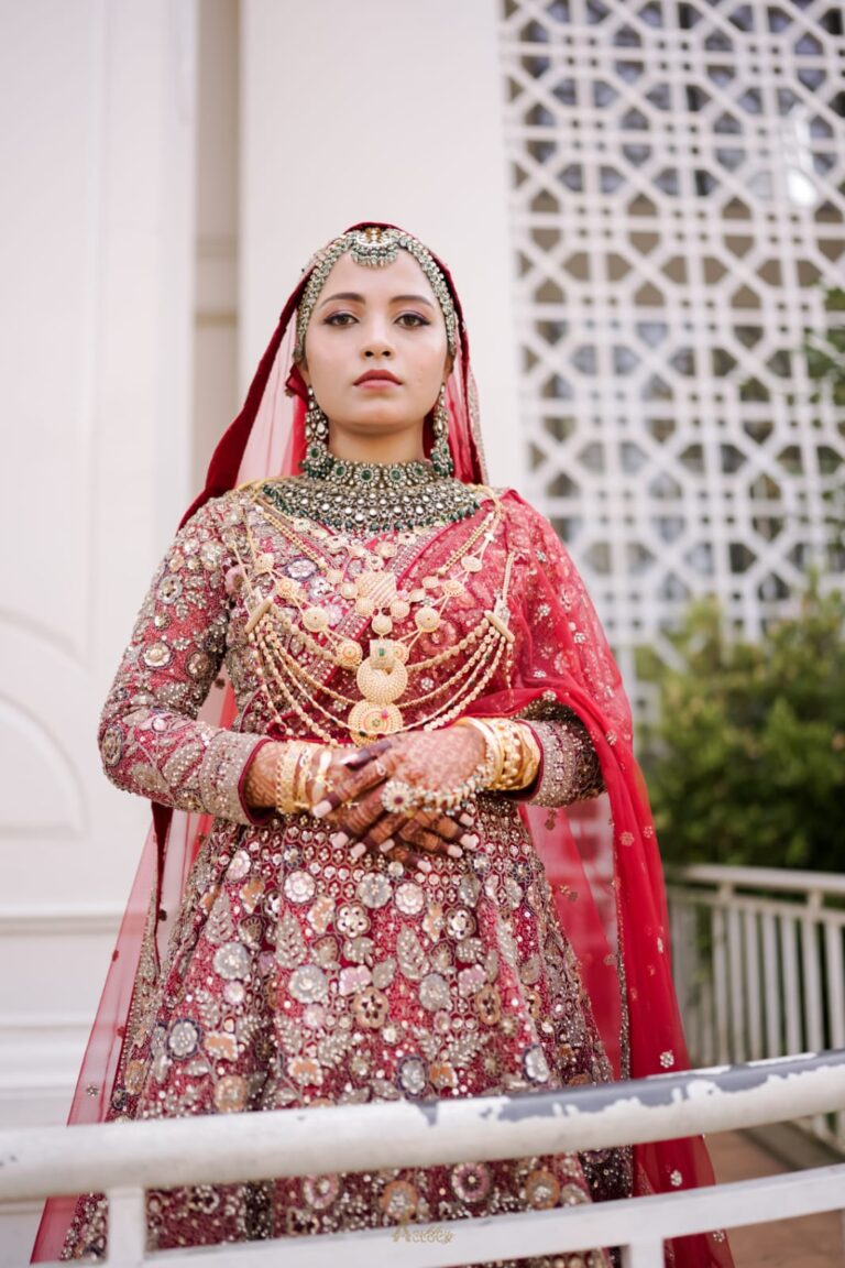 A bride in a traditional Indian wedding dress, standing outdoors with hands clasped, wearing elaborate jewelry and a red dupatta