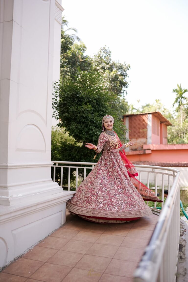 A bride in a flowing lehenga walking outdoors with her back turned, surrounded by greenery and architectural elements
