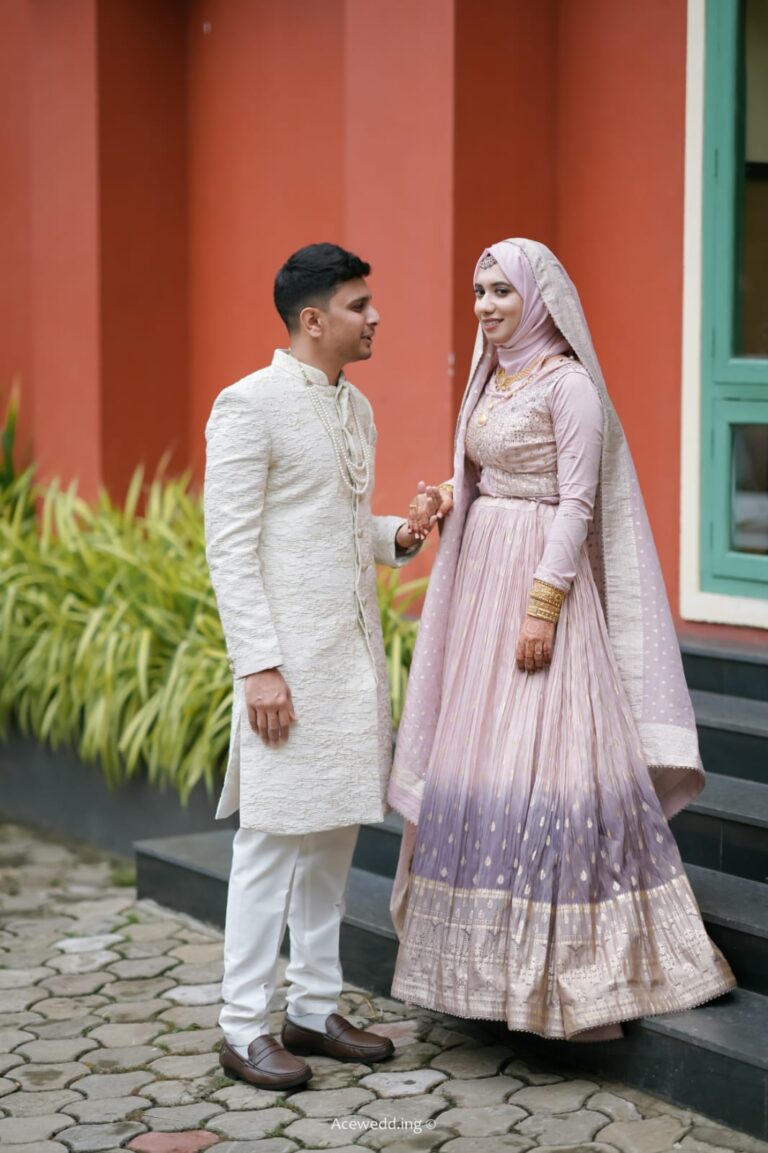 A couple in traditional light-colored wedding attire standing outdoors in front of an orange wall, facing each other and having a conversation